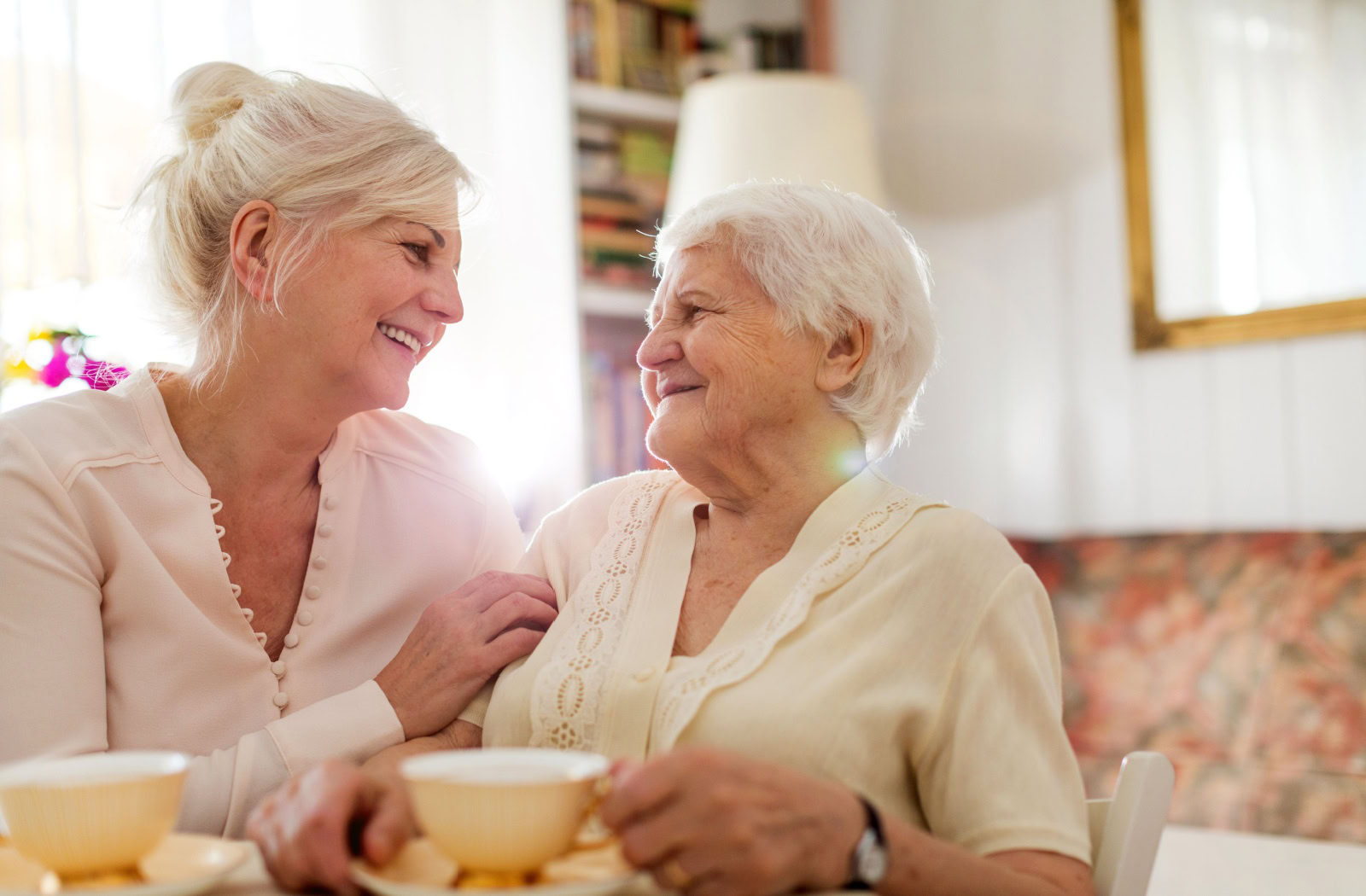 A middle-aged adult visiting their laughing parent in assisted living over a cup of coffee.