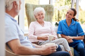 A pair of residents in assisted living sitting in the lounge and chatting happily with a smiling caregiver.