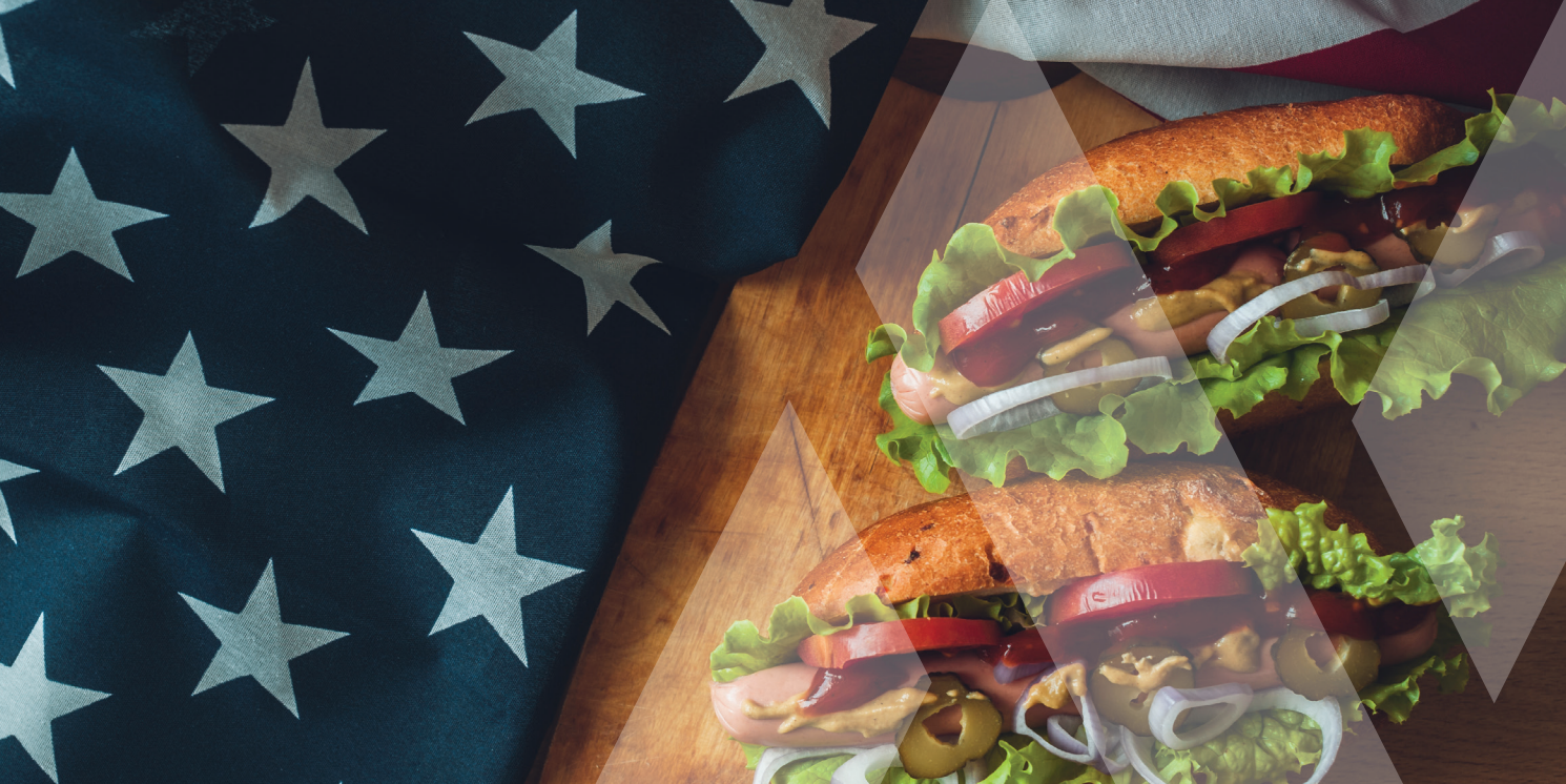 Enjoy a patriotic lunch at our senior living community with freshly made hoagie sandwiches, loaded with crisp lettuce, ripe tomato, savory onion, and black olives—served on a rustic wooden board beneath the proud backdrop of the American flag. Celebrate retirement living in comfort and style at our welcoming senior community.