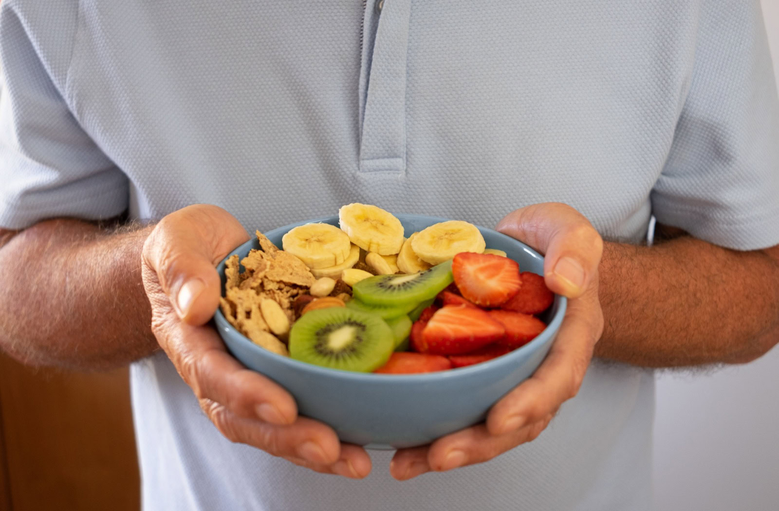 An older adult holds a bowl of grains, kiwis, strawberries, and banana slices for a healthy breakfast