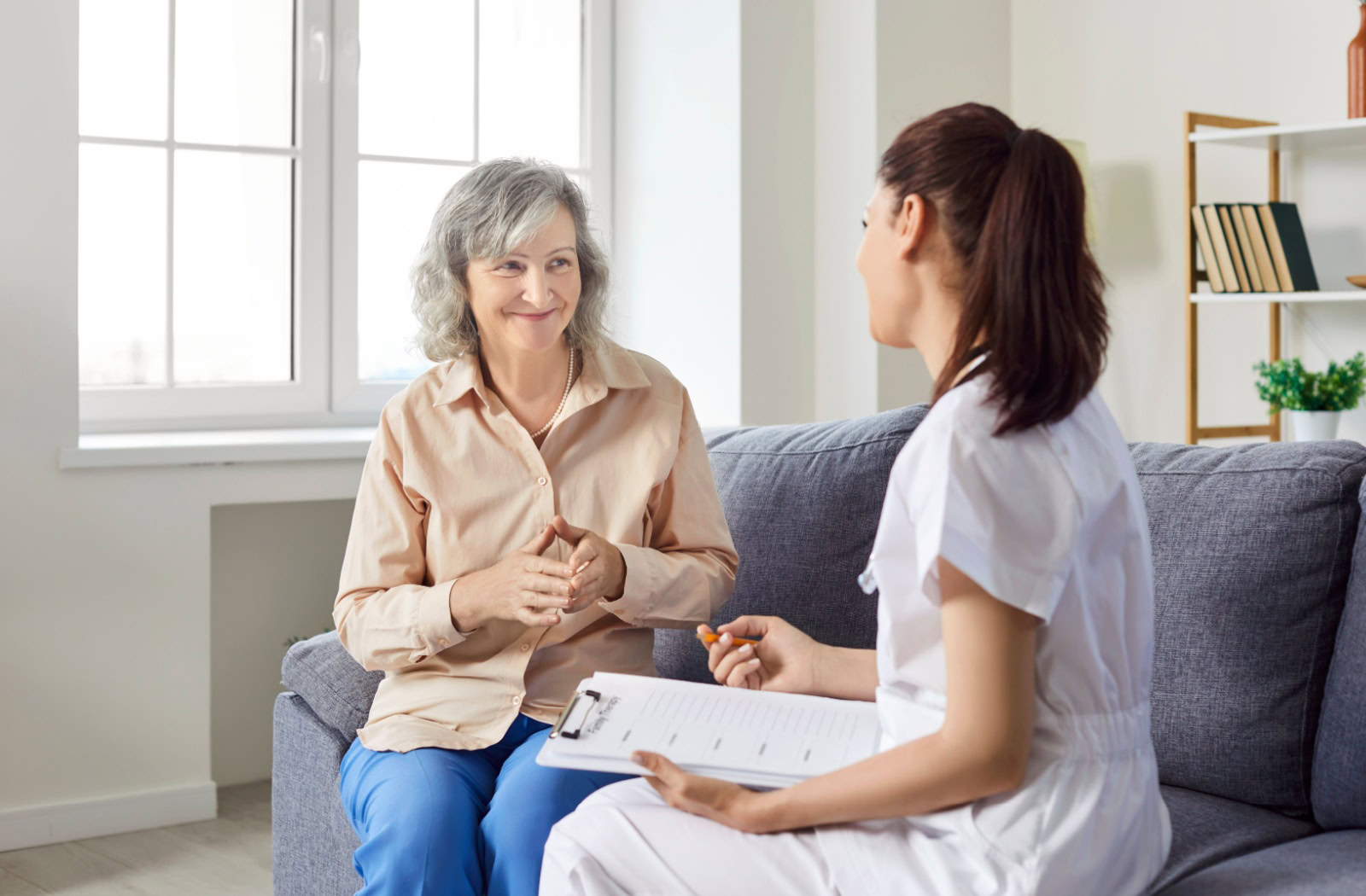 Older adult smiling during supportive conversation with healthcare professional holding clipboard on sofa