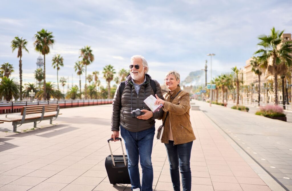 Two seniors hold a suitcase and look at a map as they walk on a boardwalk.